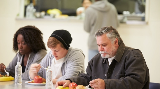 People Sitting At Table Eating Food In Homeless Shelter