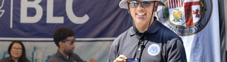Man with bucket hat and navy blue shirt holding a business card and standing in front of tent with people in the background