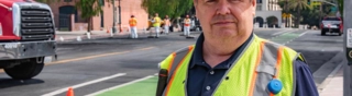 Man in white hat and yellow vest staring with buildings in the background and people working in vests
