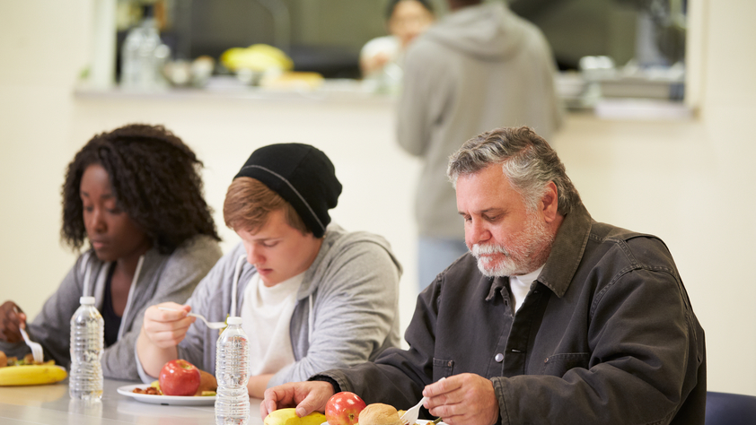 People Sitting At Table Eating Food In Homeless Shelter